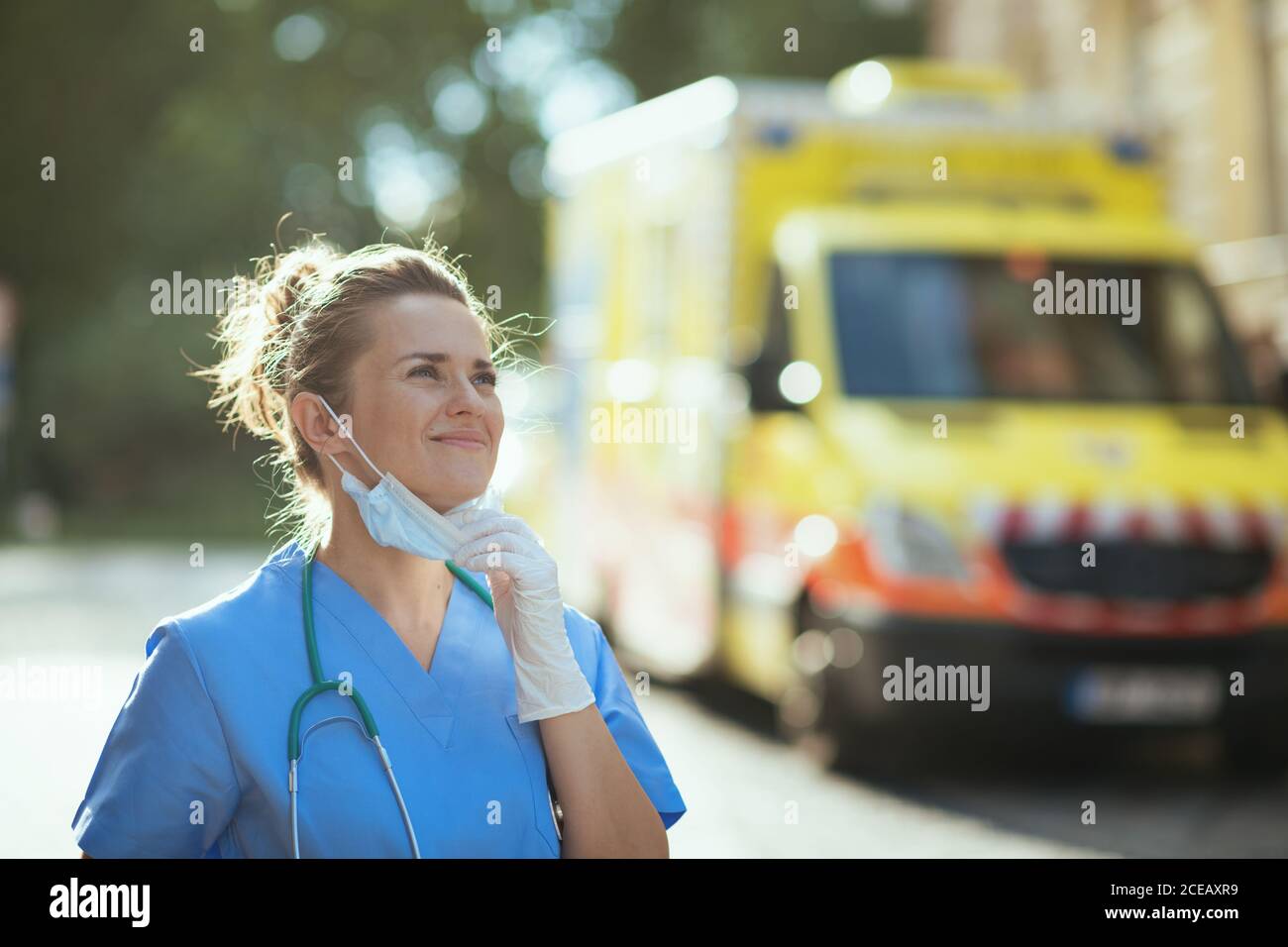 coronavirus pandemic. happy modern medical doctor woman in scrubs with stethoscope and medical mask breathing outside near ambulance. Stock Photo