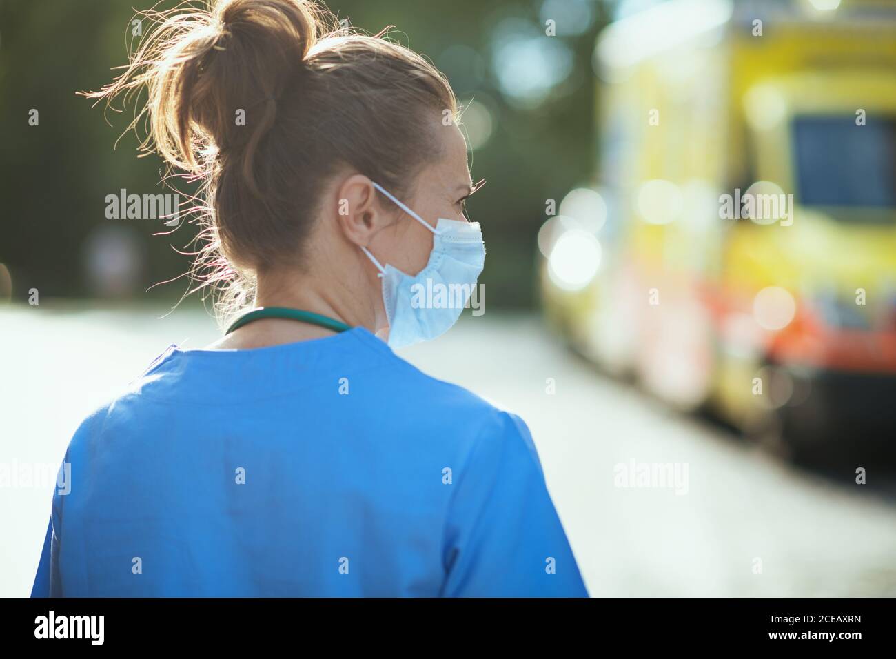 covid-19 pandemic. Seen from behind paramedic woman in uniform with medical mask outdoors near ambulance. Stock Photo