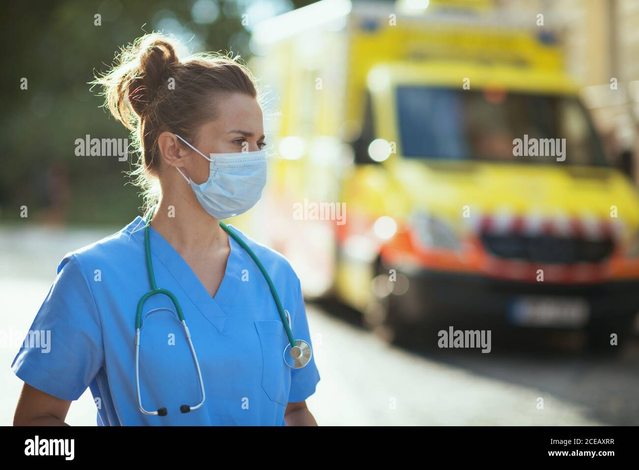 covid-19 pandemic. pensive modern medical doctor woman in uniform with stethoscope and medical mask outside near ambulance. Stock Photo