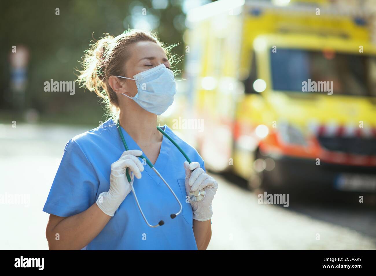coronavirus pandemic. tired modern paramedic woman in uniform with stethoscope and medical mask breathing outside near ambulance. Stock Photo