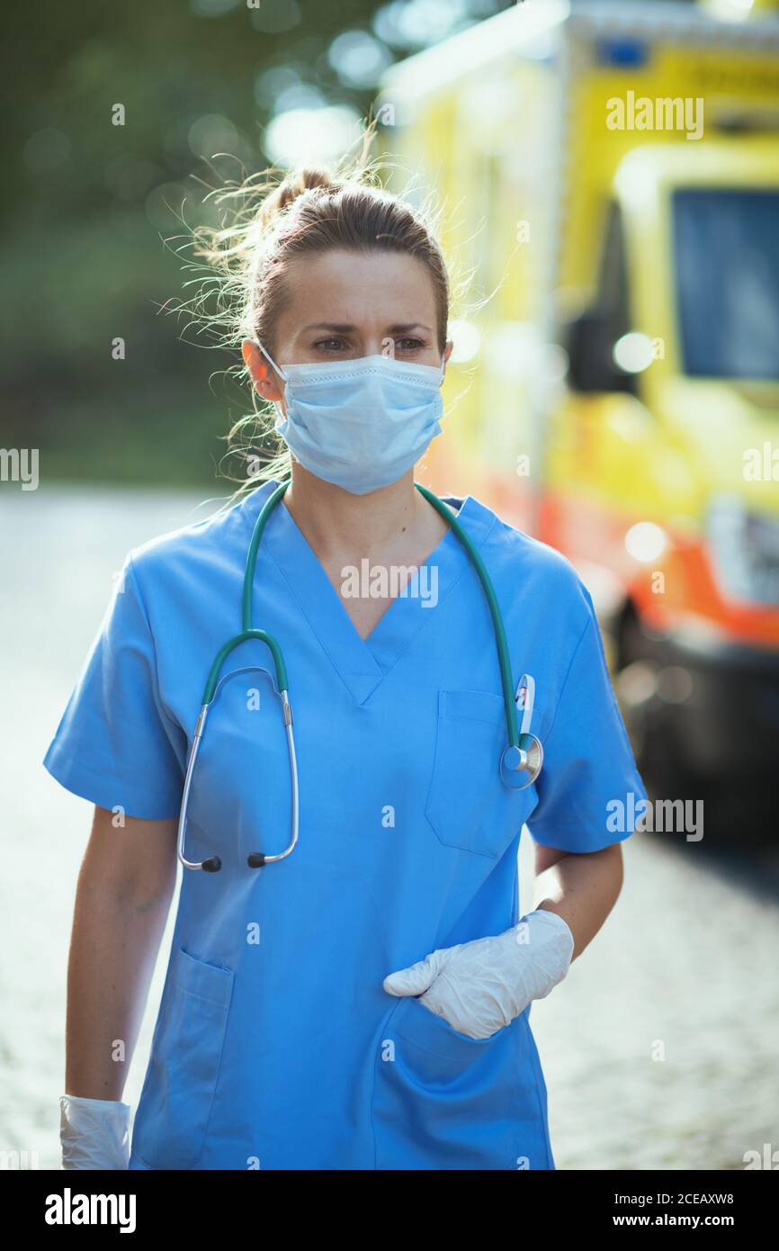 coronavirus pandemic. modern paramedic woman in scrubs with stethoscope and medical mask outdoors near ambulance. Stock Photo