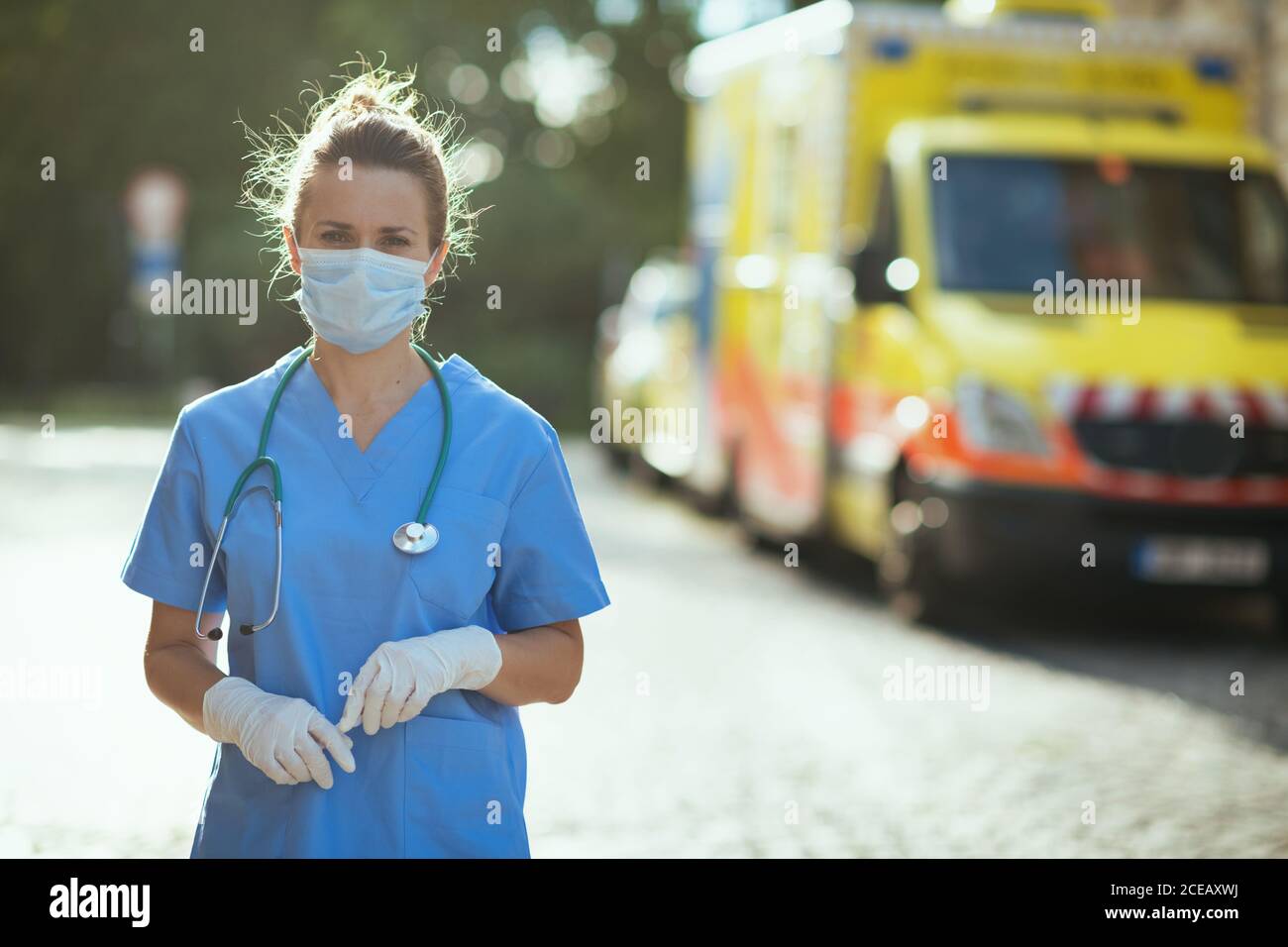covid-19 pandemic. Portrait of modern medical doctor woman in uniform with stethoscope and medical mask outdoors near ambulance. Stock Photo
