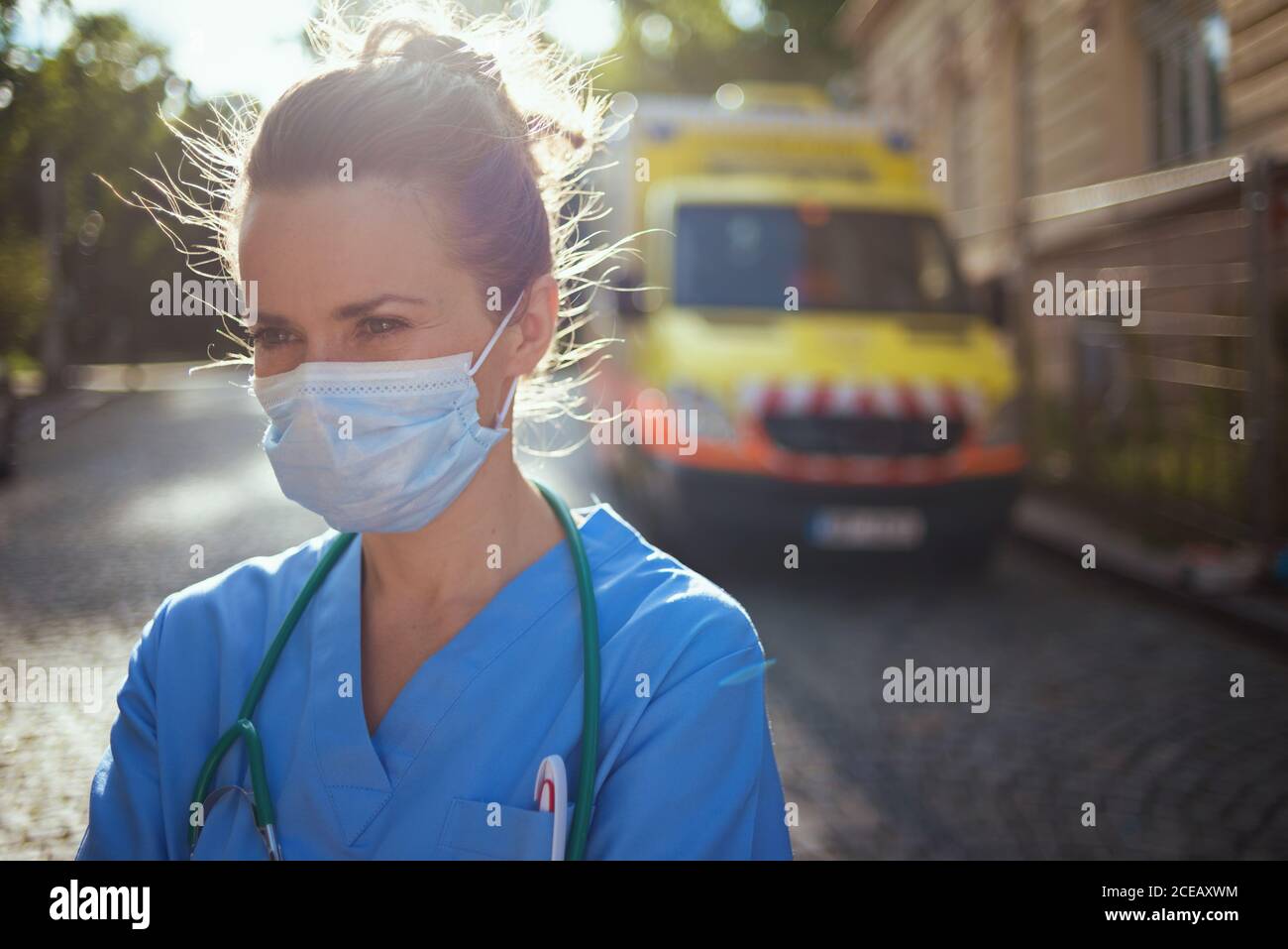 covid-19 pandemic. pensive modern paramedic woman in scrubs with stethoscope and medical mask outside near ambulance. Stock Photo