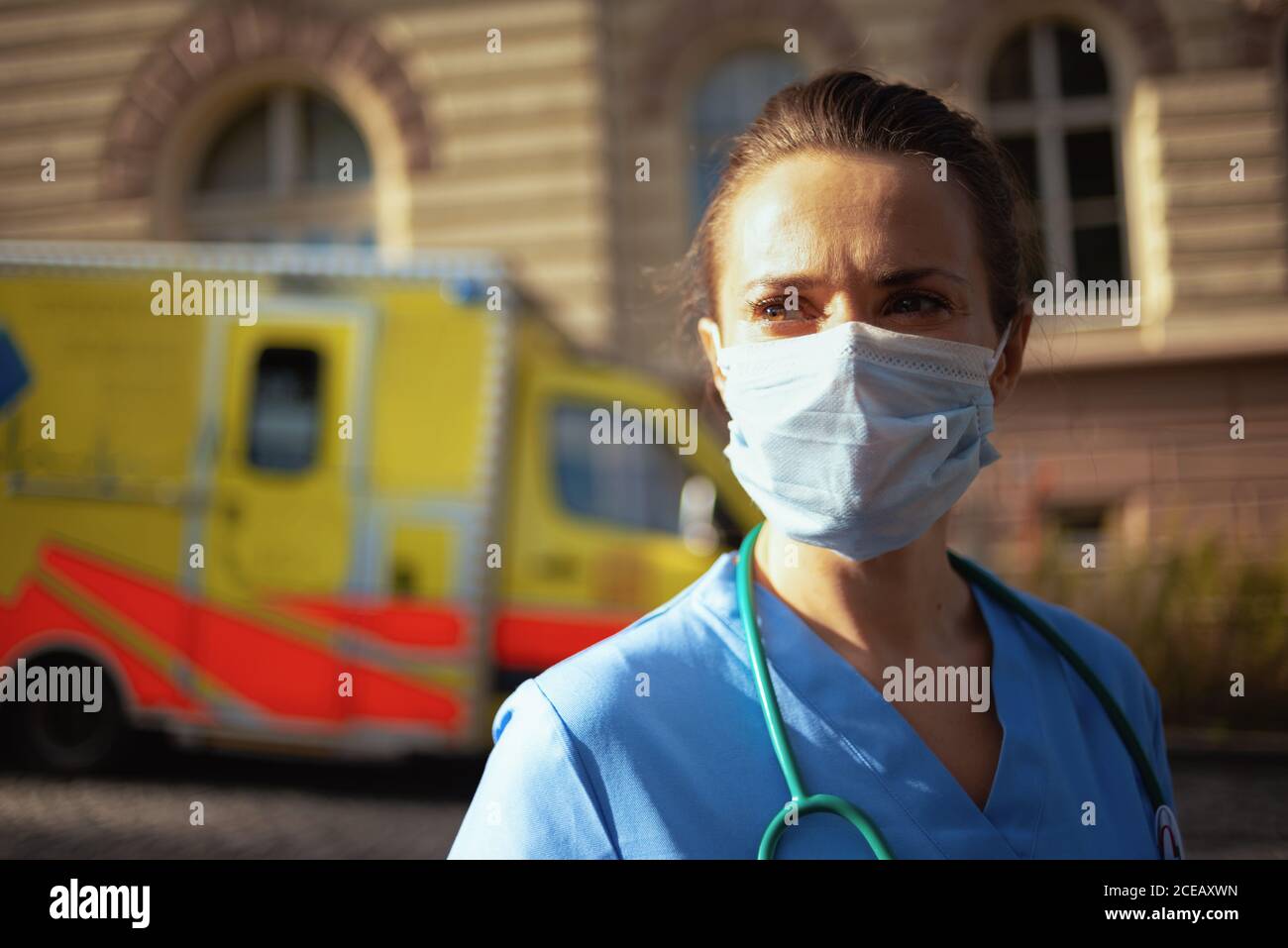 covid-19 pandemic. pensive modern medical doctor woman in scrubs with stethoscope and medical mask outside near ambulance. Stock Photo