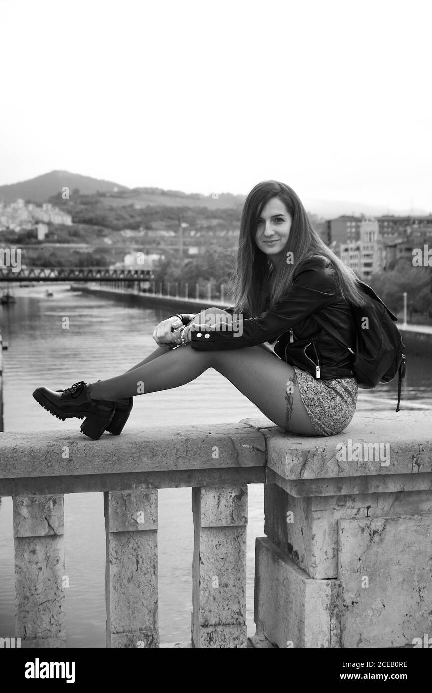 Side view of young attractive Woman in leather jacket and short skirt with tattoo on leg and backpack sitting on stone parapet of bridge in Bilbao and looking at camera on background of river Stock Photo