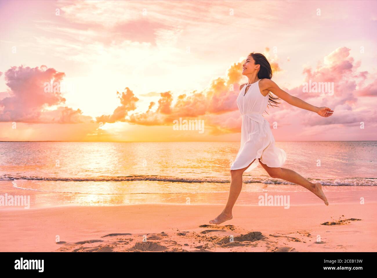Freedom wellness well-being happiness concept. Happy carefree Asian woman feeling blissful jumping of joy on peaceful beach at sunset. Serenity Stock Photo