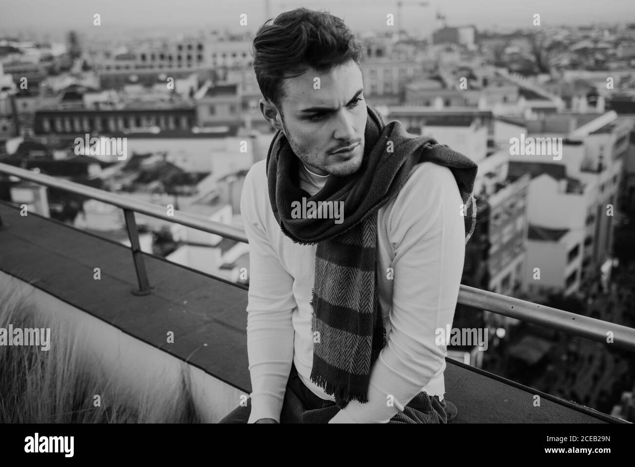 Black and white young stylish guy in casual wear with scarf sitting on roof and looking away on cityscape background in Madrid, Spain Stock Photo
