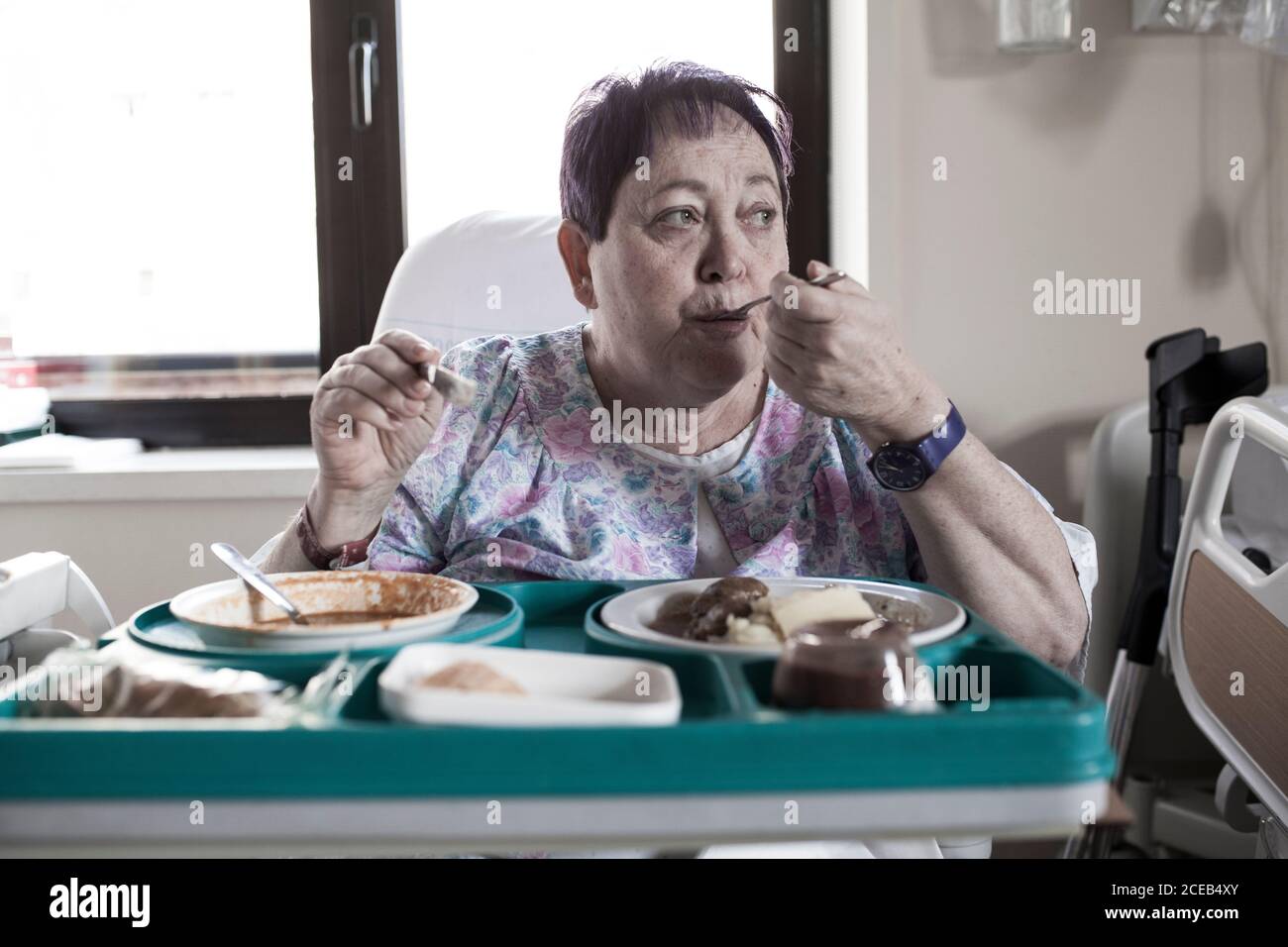 Mature woMan hospitalized for knee surgery, sitting in the chair, eating food from the tray Stock Photo