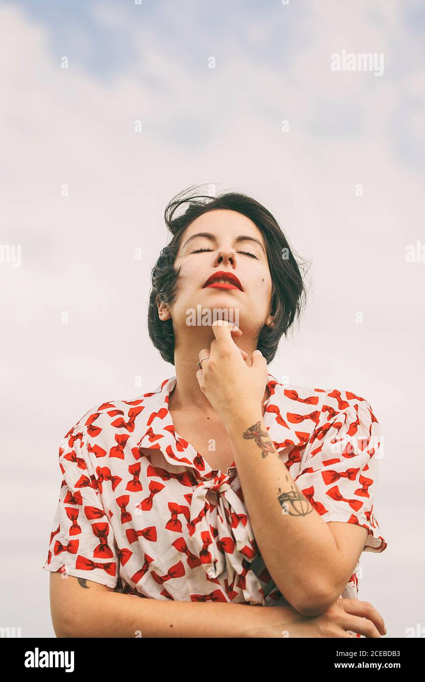 From below view of attractive brunette Woman in patterned shirt with tattoos on arm standing on background of cloudy sky with closed eyes Stock Photo