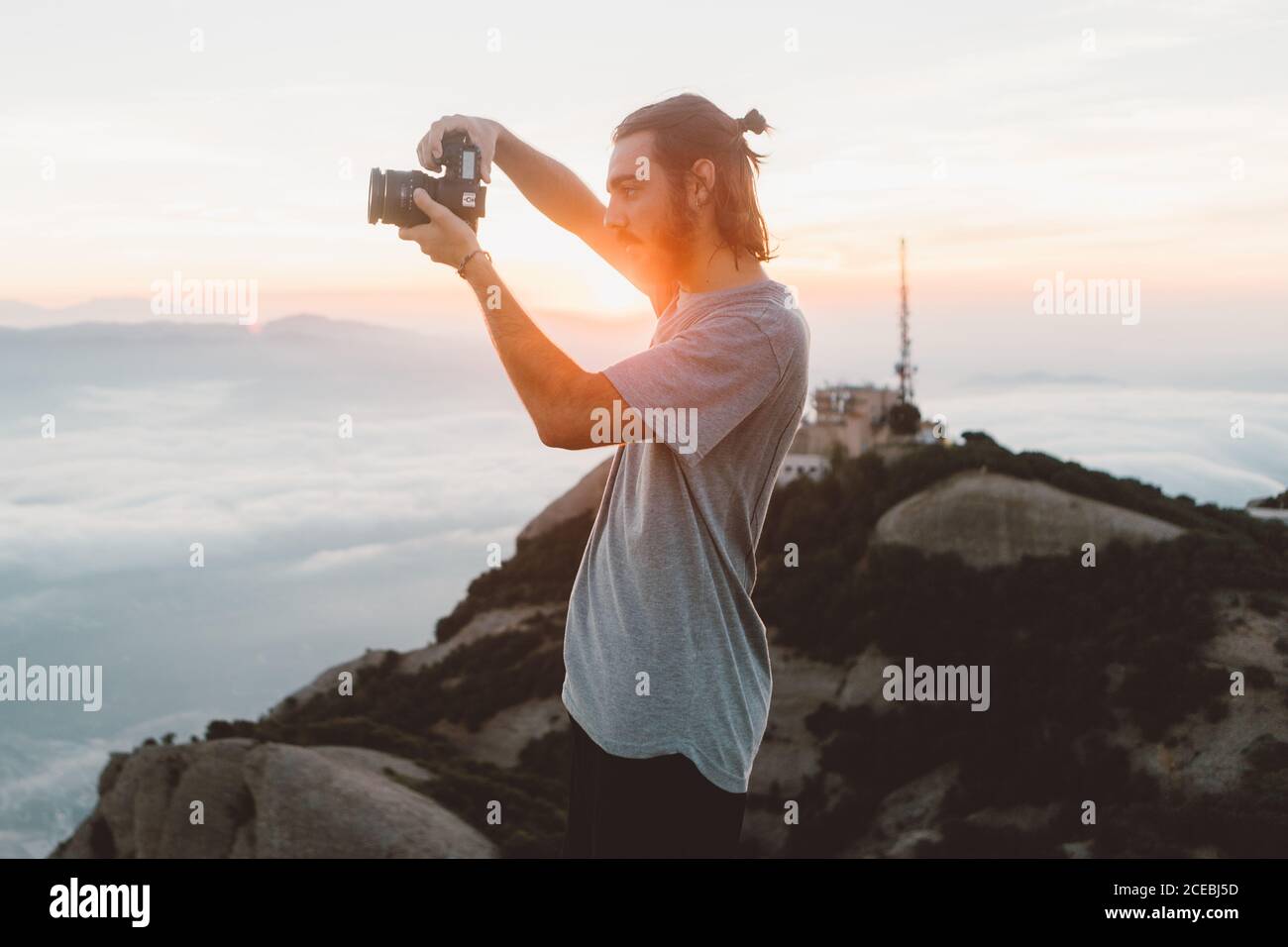 Side view of handsome bearded guy using professional photo camera to take photos on amazing nature while standing on mountain during sunrise in Barcelona, Spain Stock Photo