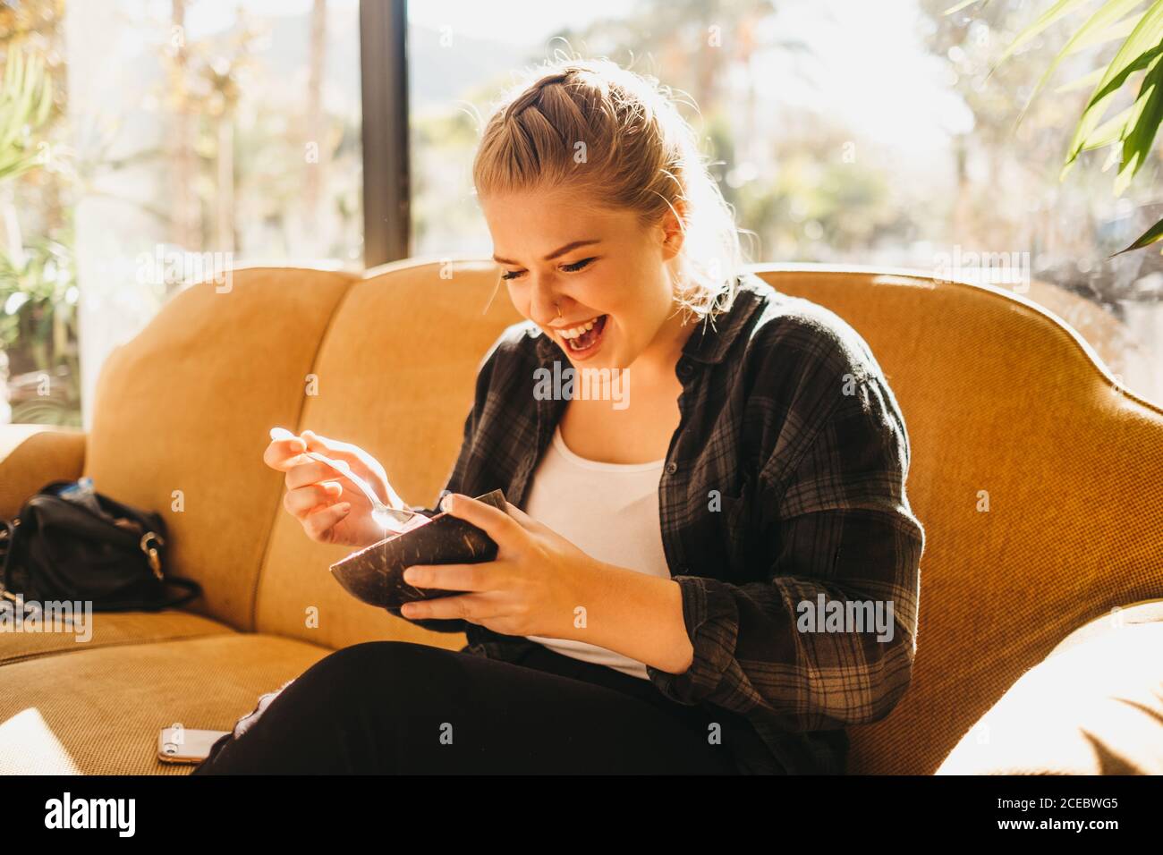 Pretty young female with bowl and spoon smiling while sitting on comfortable couch near window Stock Photo