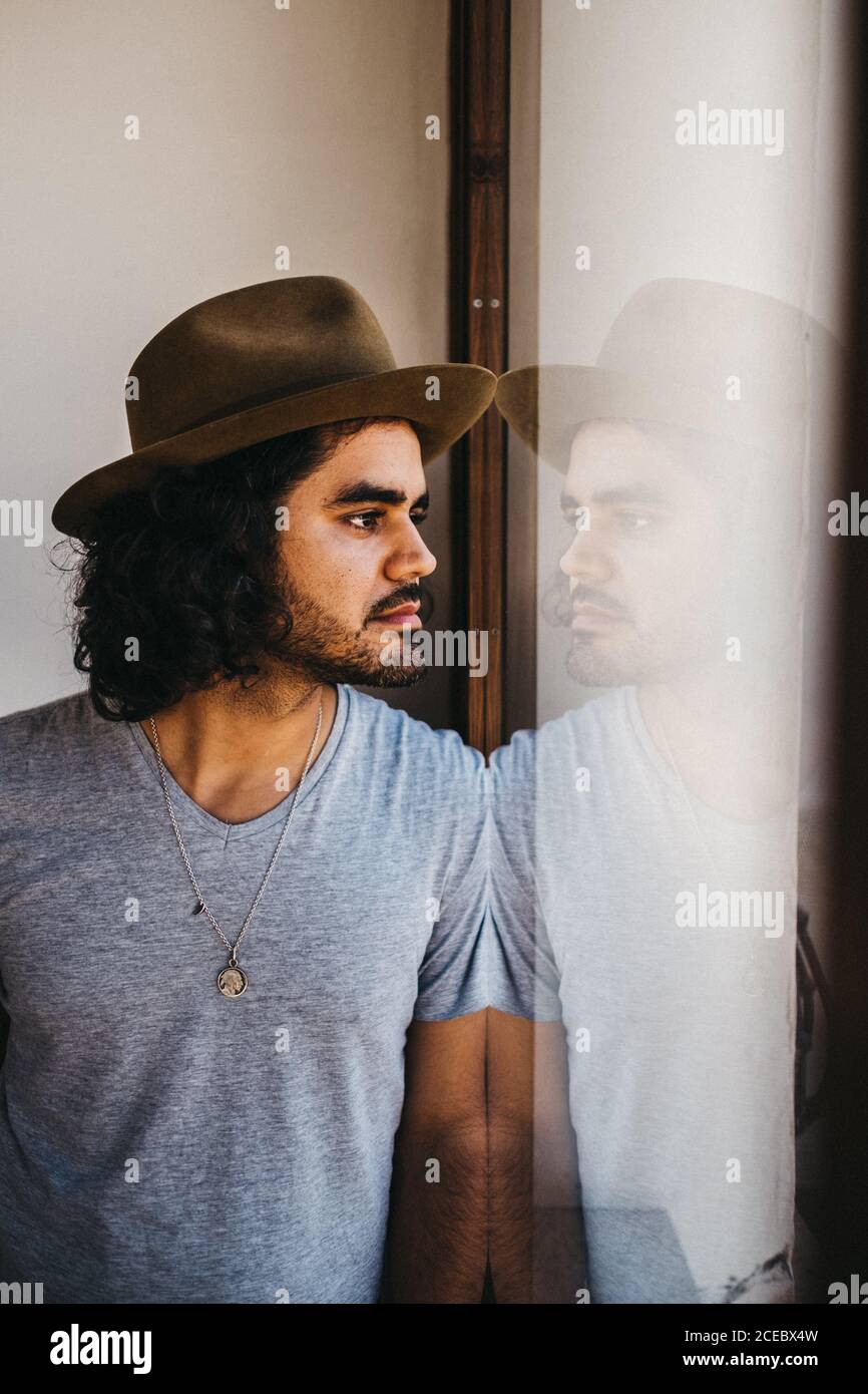 Adult modern man with curly hair and beard wearing hat and looking pensively in window Stock Photo