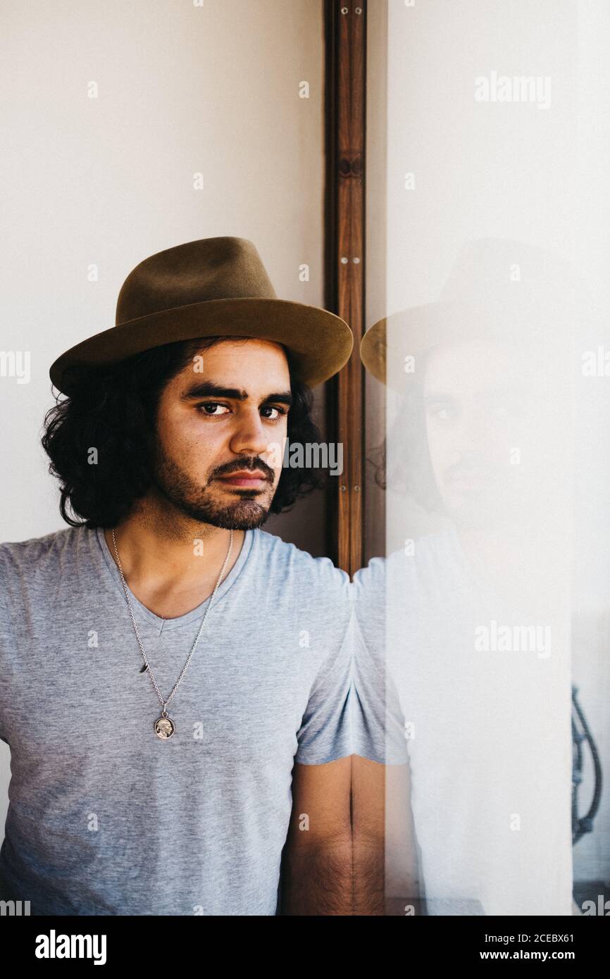 Adult modern man with curly hair and beard wearing hat and looking pensively in window Stock Photo