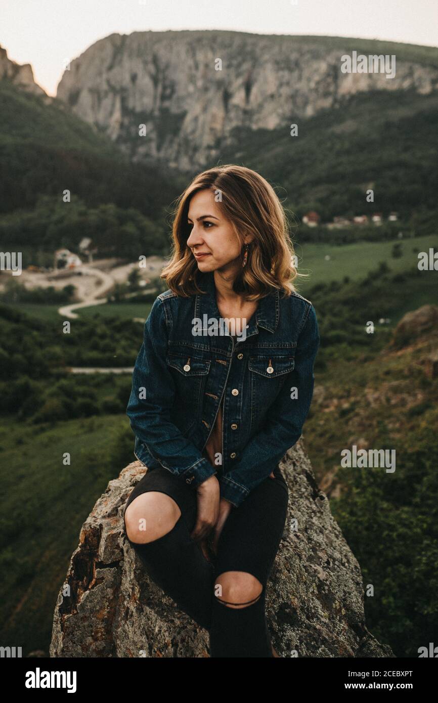 Lovely young female smiling and looking away while sitting on rock on background of picturesque countryside Stock Photo