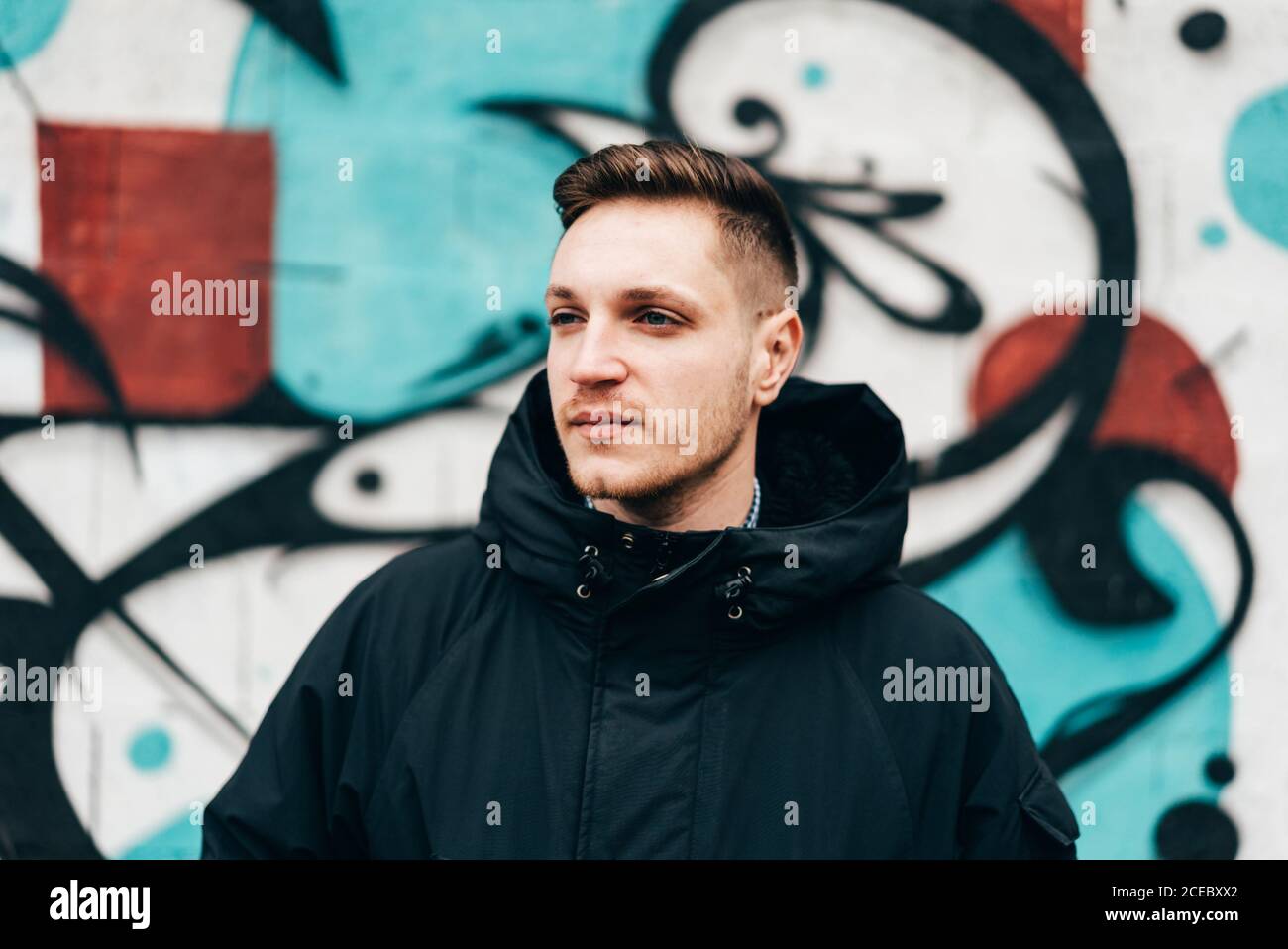 Thoughtful young man standing at graffiti wall with hands in pockets. Stock Photo