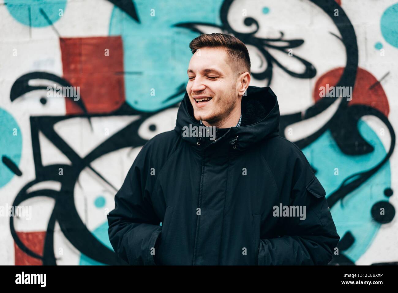 Thoughtful young man standing at graffiti wall with hands in pockets. Stock Photo
