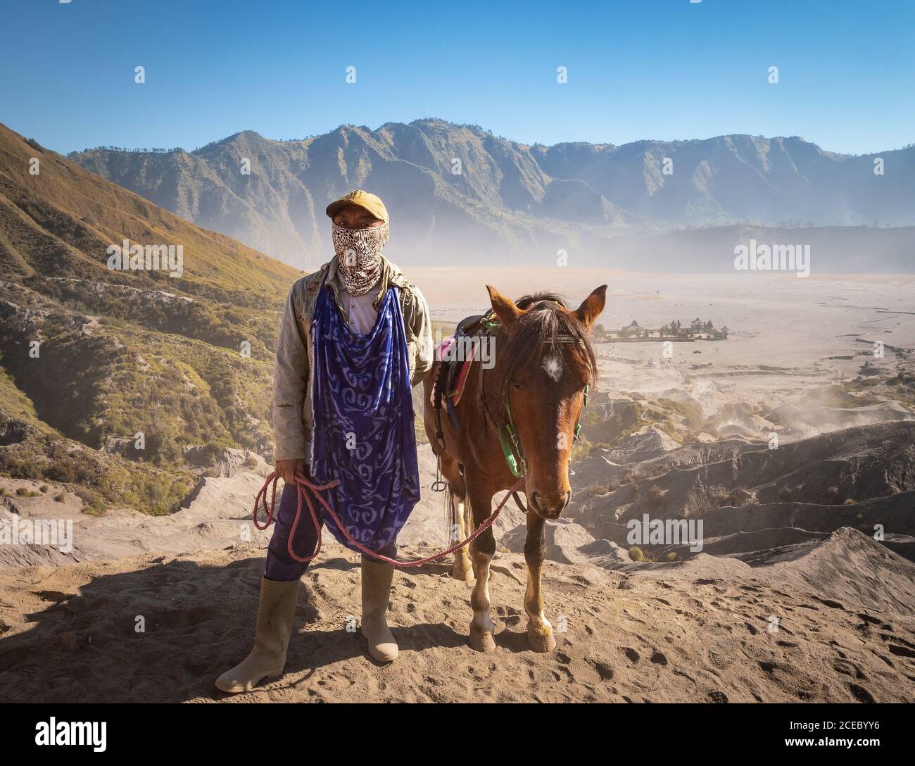 Java Island, Indonesia - August, 14 2015: Ethnic person with scarf on face looking at camera while standing near lovely horse not far from Mount Bromo on sunny day Stock Photo