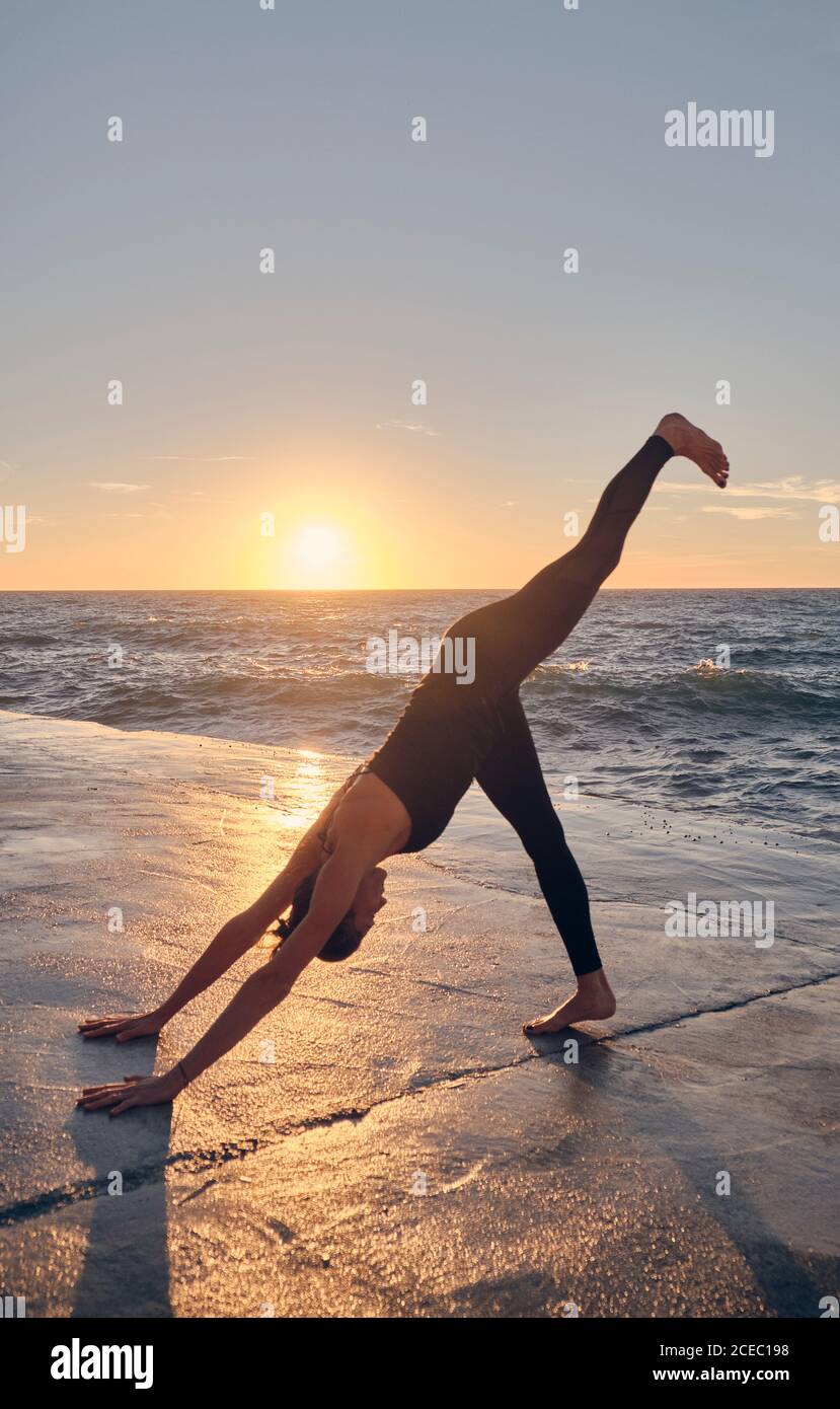 Side view of young female in sportswear standing in Downward Facing Dog Pose on stone surface against waving sea and wonderful sunset sky Stock Photo