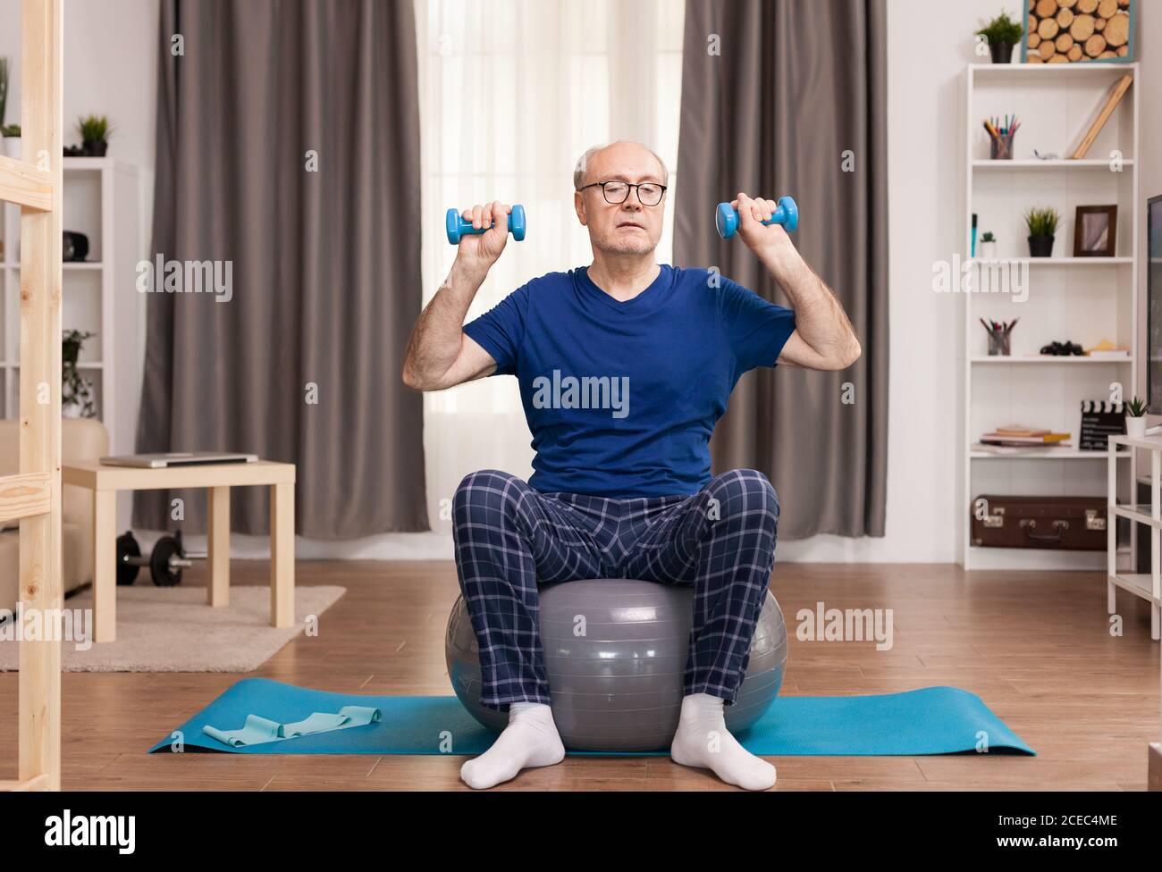 Senior man with vitality exercising in living room. Old person pensioner online internet exercise training at home sport activity with dumbbell, resistance band, swiss ball at elderly retirement age. Stock Photo