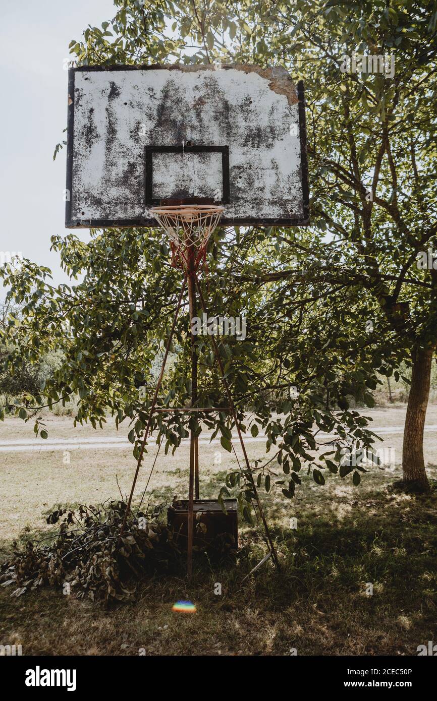 Old basketball hoop with broken board standing on the grass in shadow under the tree with backlighting sun near the gravel road of the Serbian village Stock Photo