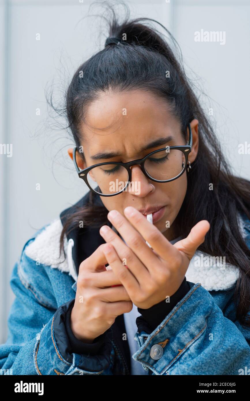 Attractive female in glasses lighting cigarette and covering flame with hand while smoking on city street on windy day Stock Photo