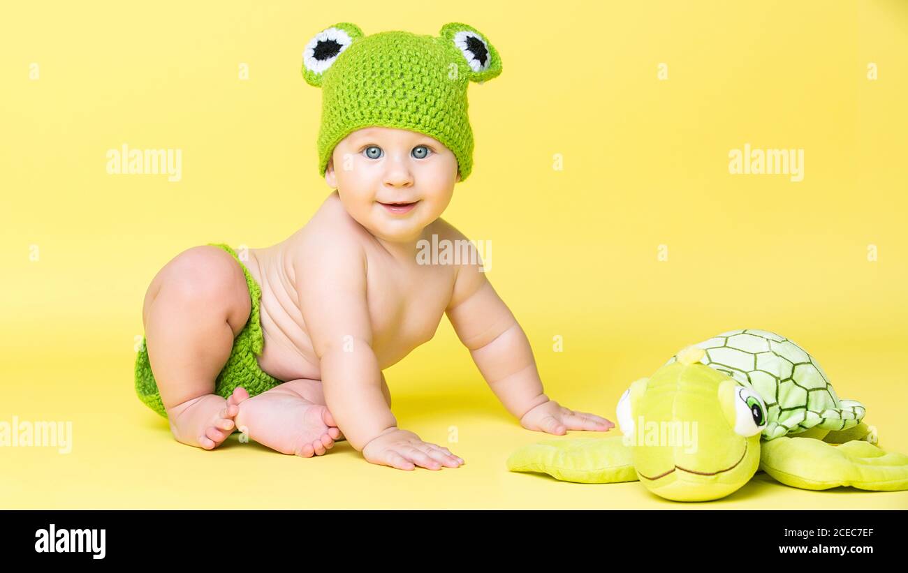 Adorable little boy in frog hat sitting at turtle toy on yellow background. Stock Photo