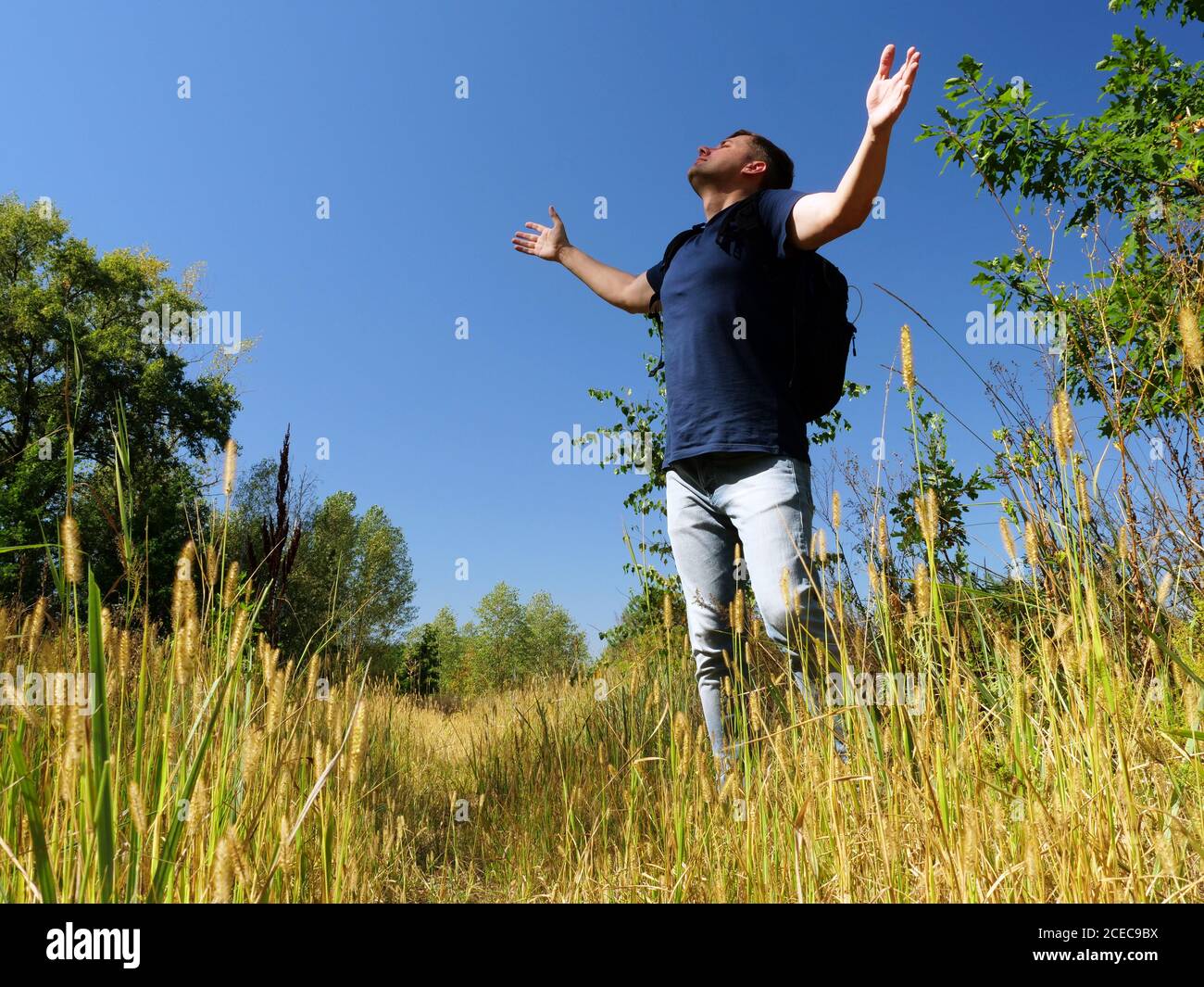 A hiker with a backpack stretches out his hands to the summer sky in nature. Active rest and relaxation. Stock Photo