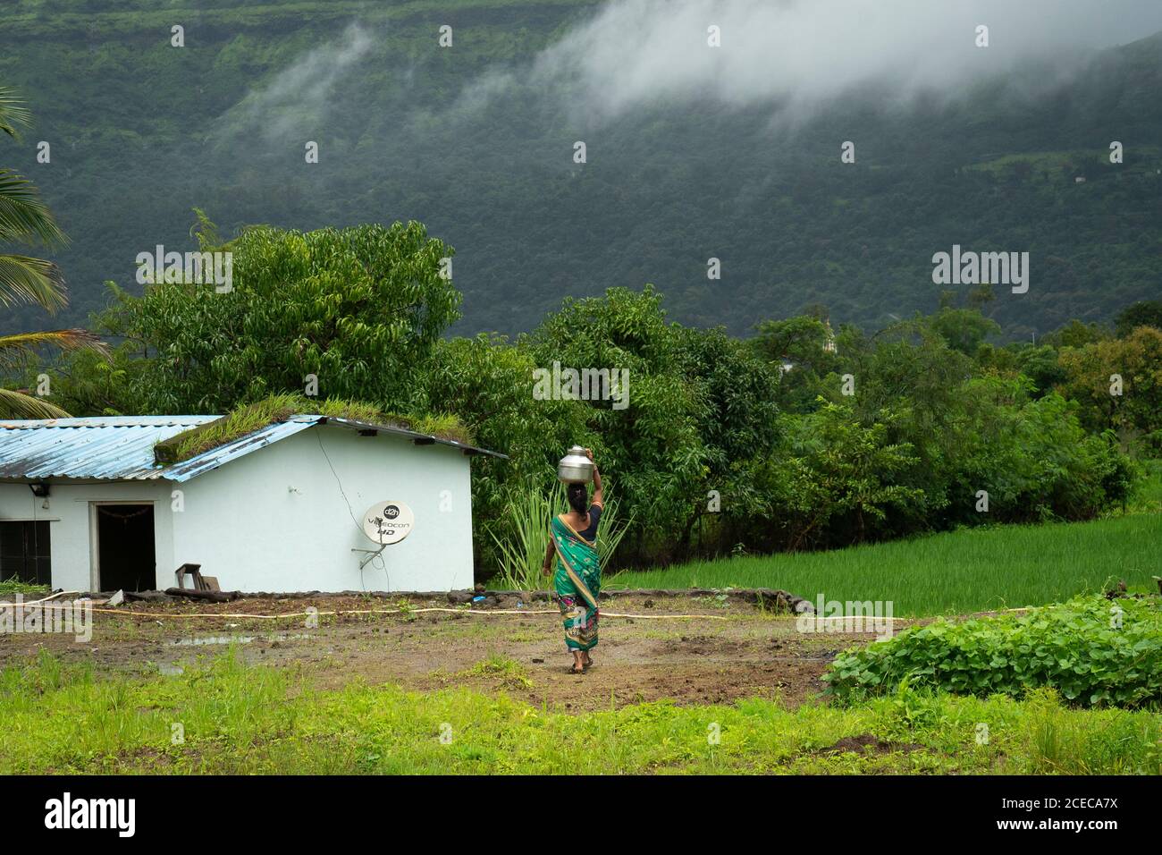 a lady villager walking with a water pot on her head with the background of Sahyadri mountain with floating clouds. Stock Photo
