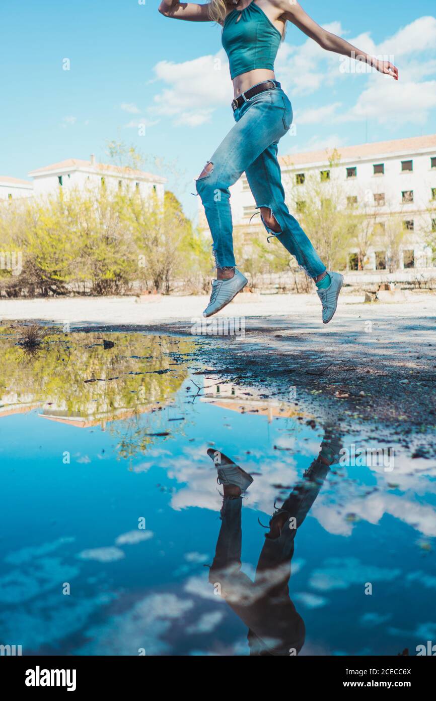 faceless shot of slim girl in denim jumping high above puddle reflecting blue sky and city. Stock Photo