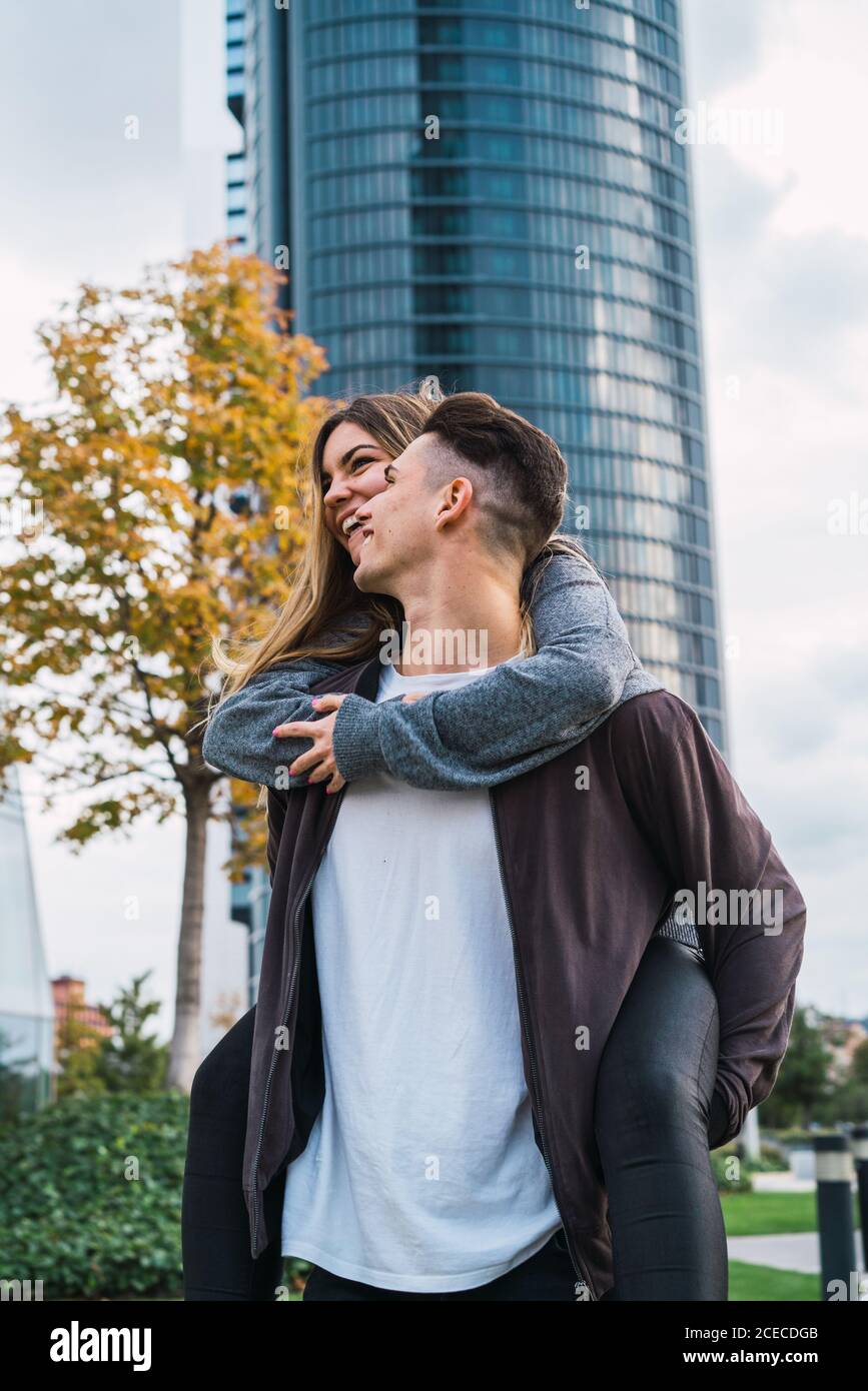 Young man carrying charming girlfriend on back while posing on urban background. Stock Photo