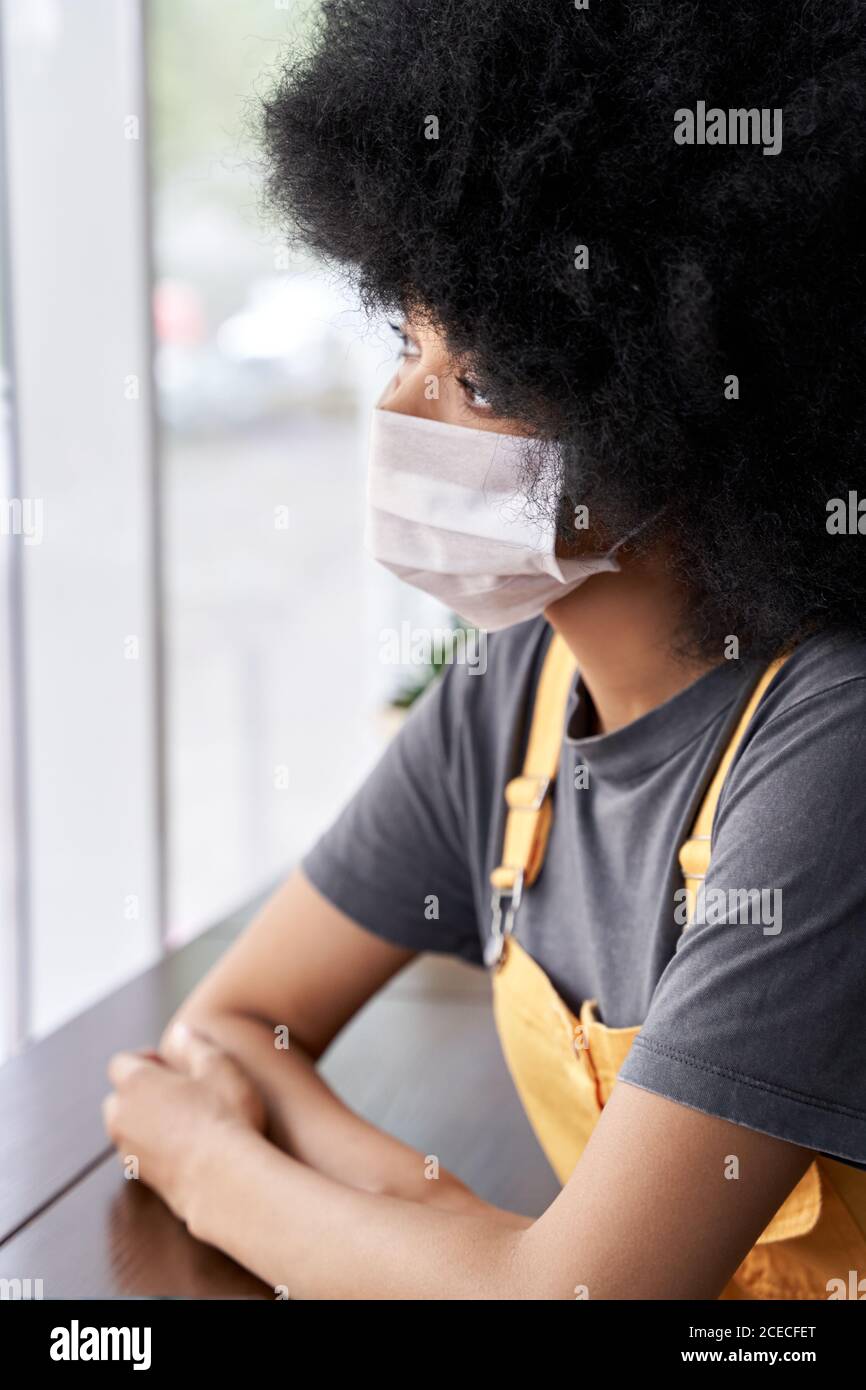 Thoughtful pensive African lady wearing face mask looking through window. Stock Photo