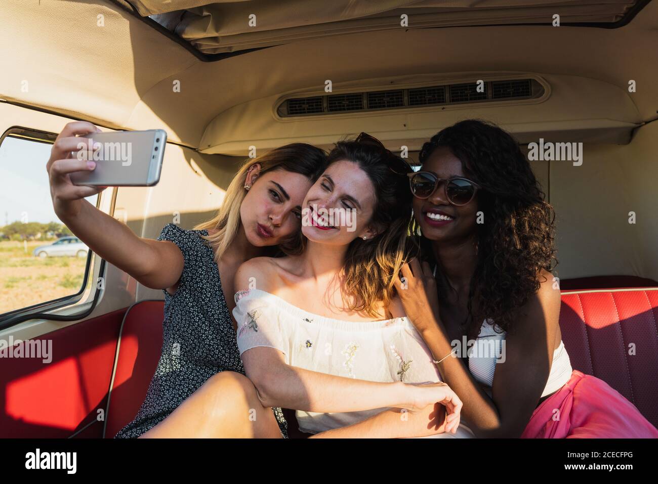 Three cheerful young women sitting inside vintage van and posing for selfie while traveling together Stock Photo