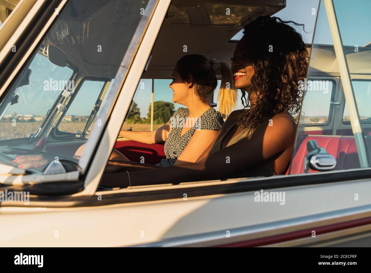 Side view of two young women sitting inside vintage van and smiling while traveling in nature together Stock Photo