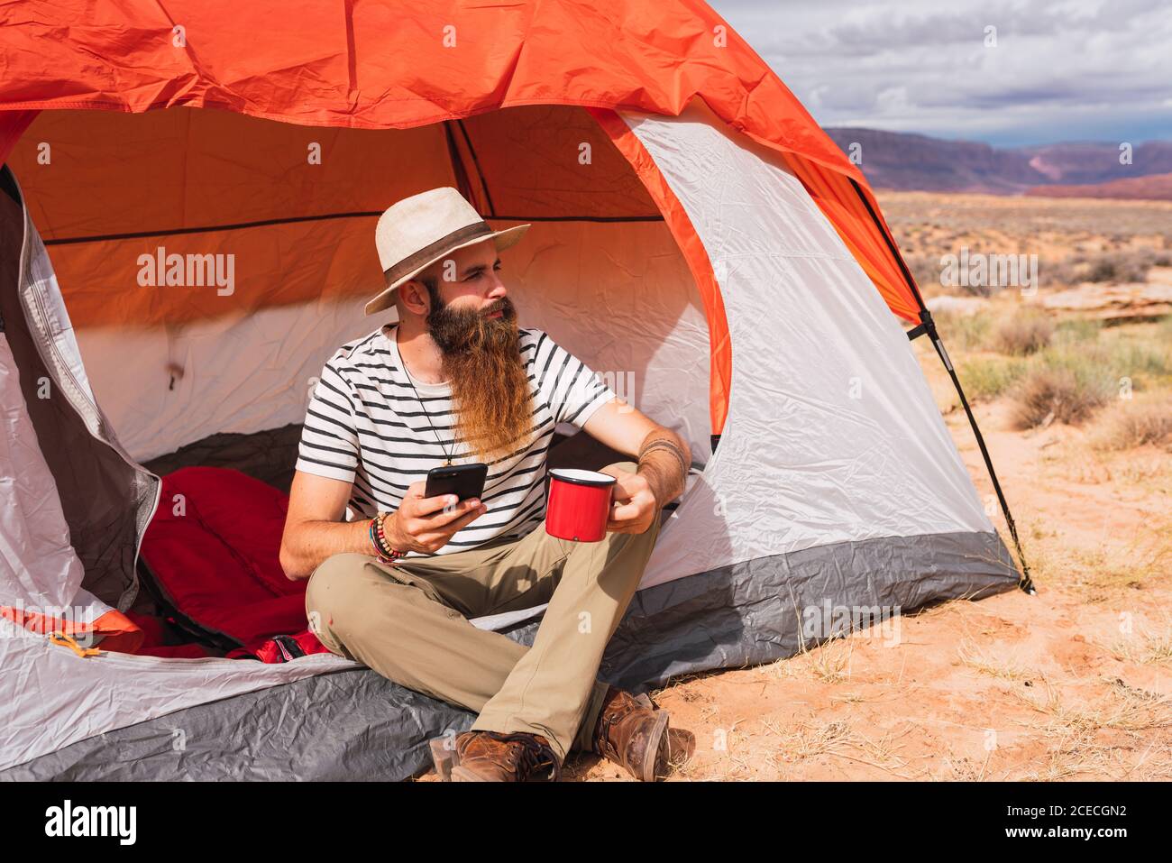 Bearded guy in casual outfit holding mug of hot beverage and modern smartphone while sitting near tent and looking away in beautiful nature Stock Photo