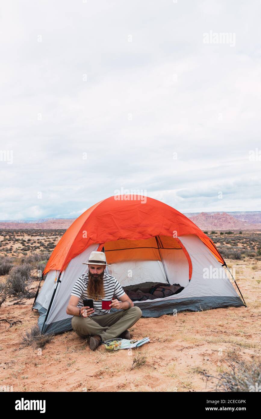 Bearded guy in casual outfit holding mug of hot beverage, salad and modern smartphone while sitting near tent and looking away in beautiful nature Stock Photo