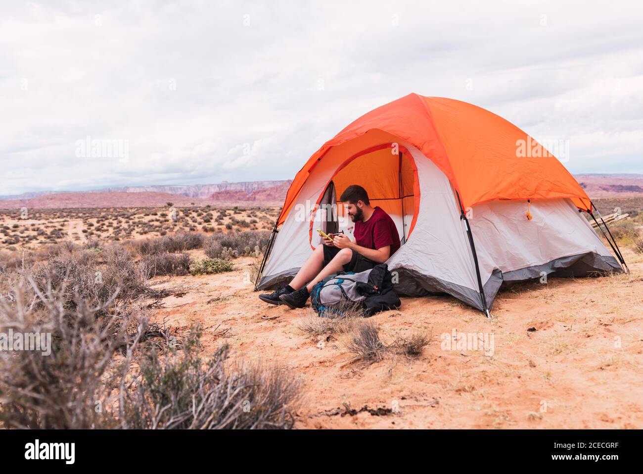 Bearded guy in casual outfit holding mug of hot beverage and modern smartphone while sitting near tent and looking away in beautiful nature Stock Photo