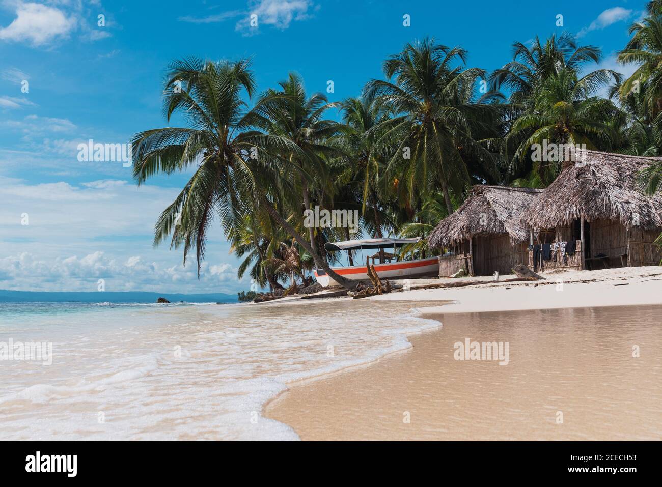 Picturesque view of huts, boat and tropical forest on sand beach near sea and blue sky in San Blas islands, Panama Stock Photo