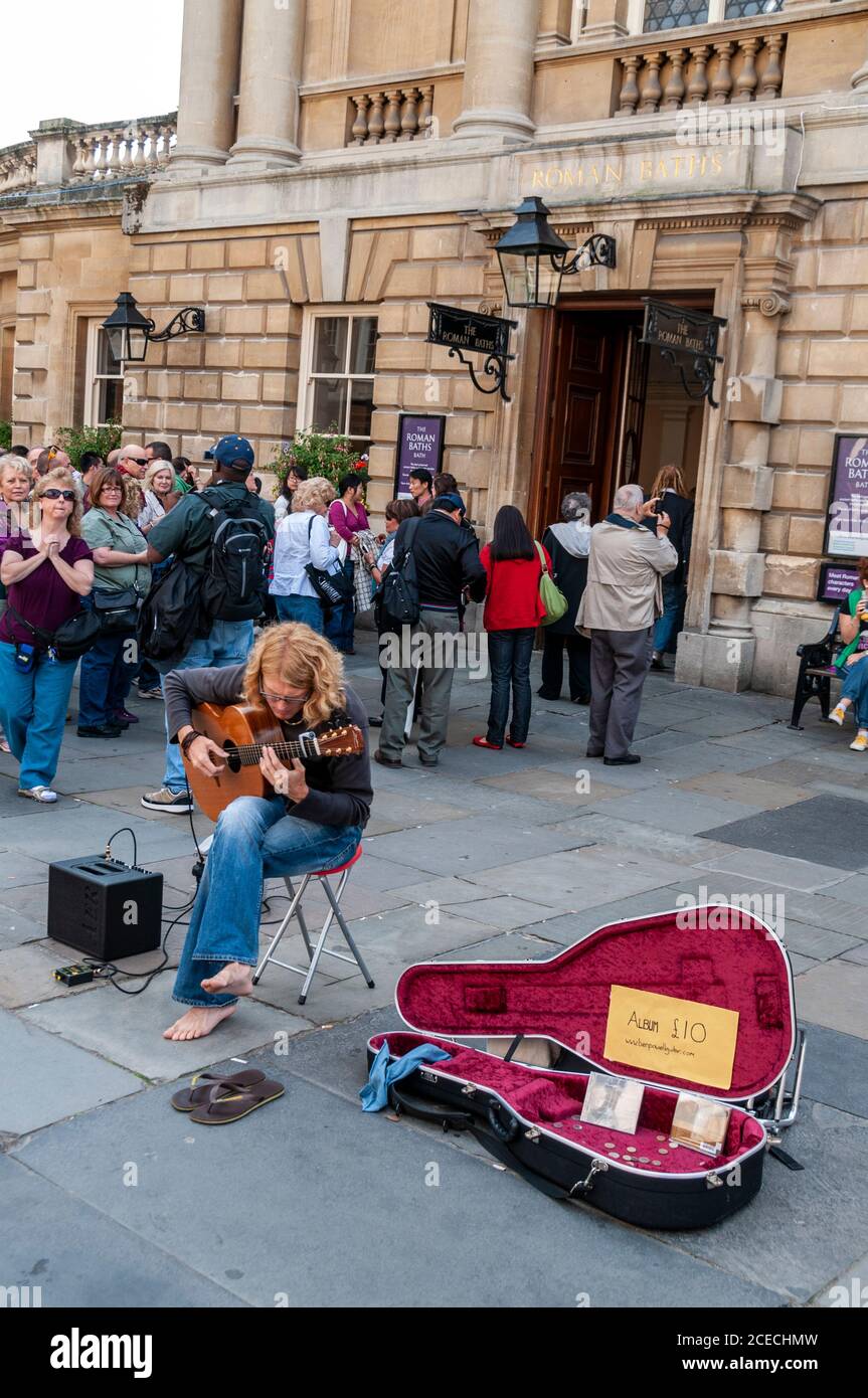 A street busker with his guitar playing to visitors queuing outside the Roman Baths in Abbey Churchyard in the Roman city of Bath in Britain. Stock Photo