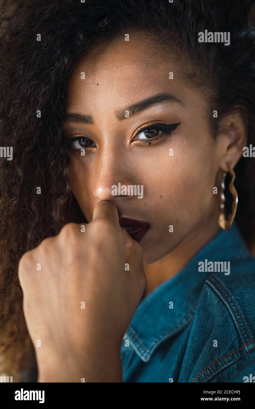 Young attractive African-American Woman with bright makeup and dark curly hair in denim shirt looking at camera with hand on lips Stock Photo