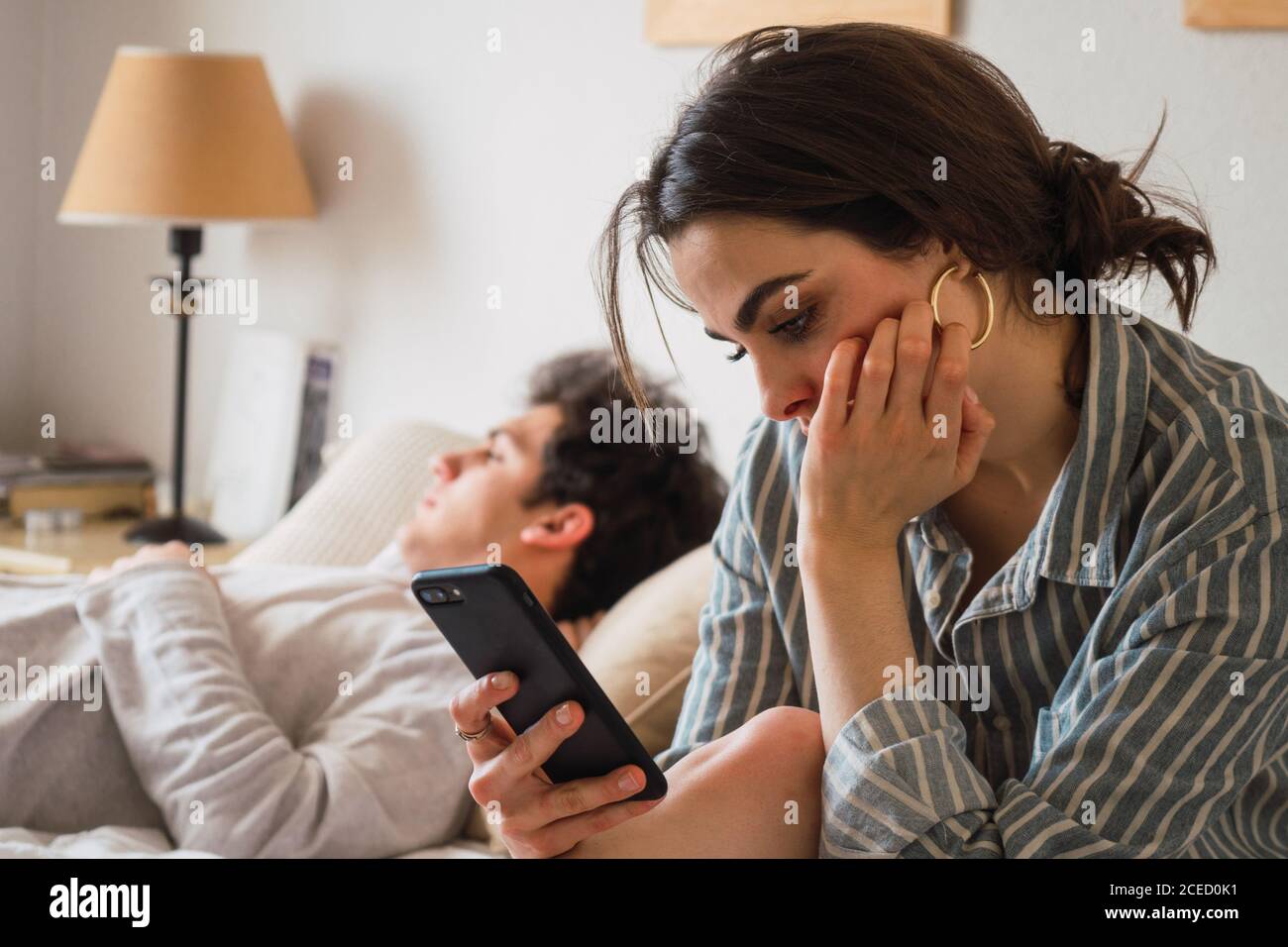 Side view of young sad thoughtful Woman sitting and reading smartphone while boyfriend sleeping. Stock Photo