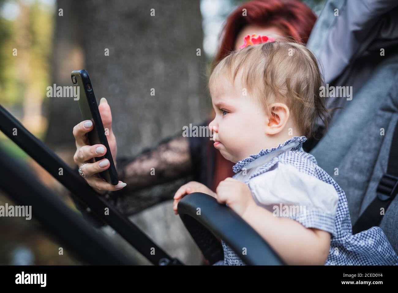 unrecognizable Woman holding modern smartphone and trying to cheer up adorable sad baby girl in stroller Stock Photo