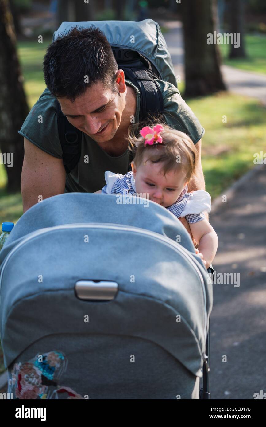 Handsome adult man smiling and putting sweet baby girl in carriage while standing on blurred background of park Stock Photo