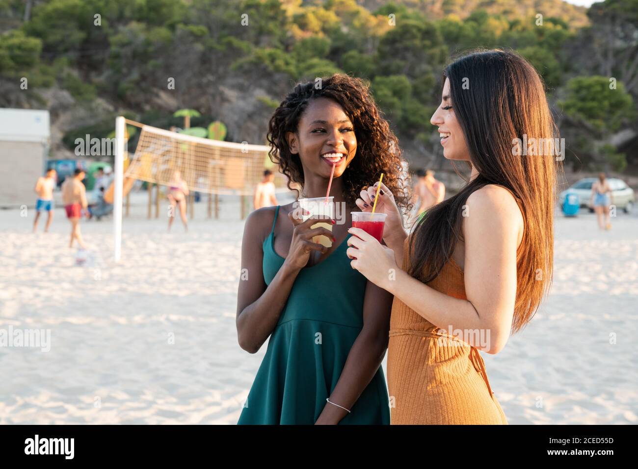 Diverse young girlfriends chilling on beach with drinks in cups and laughing while chatting in sunset light Stock Photo