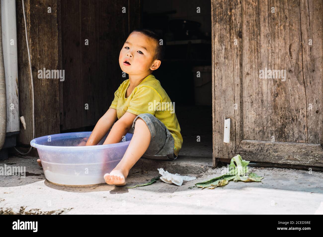 GUINZHOU, CHINA - JUNE 14, 2018: Adorable little child of Miao ethnic minority in summer clothes sitting on stone floor with hands in plastic basin with water and looking away in Guizhou province of China Stock Photo