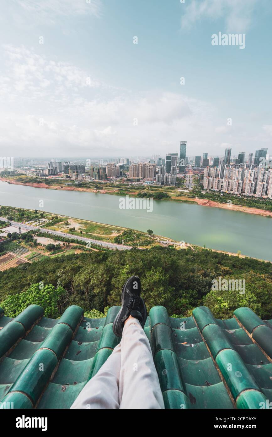 Crop legs of man chilling on roof with view of contemporary Nanning cityscape and green shore of river below, China Stock Photo