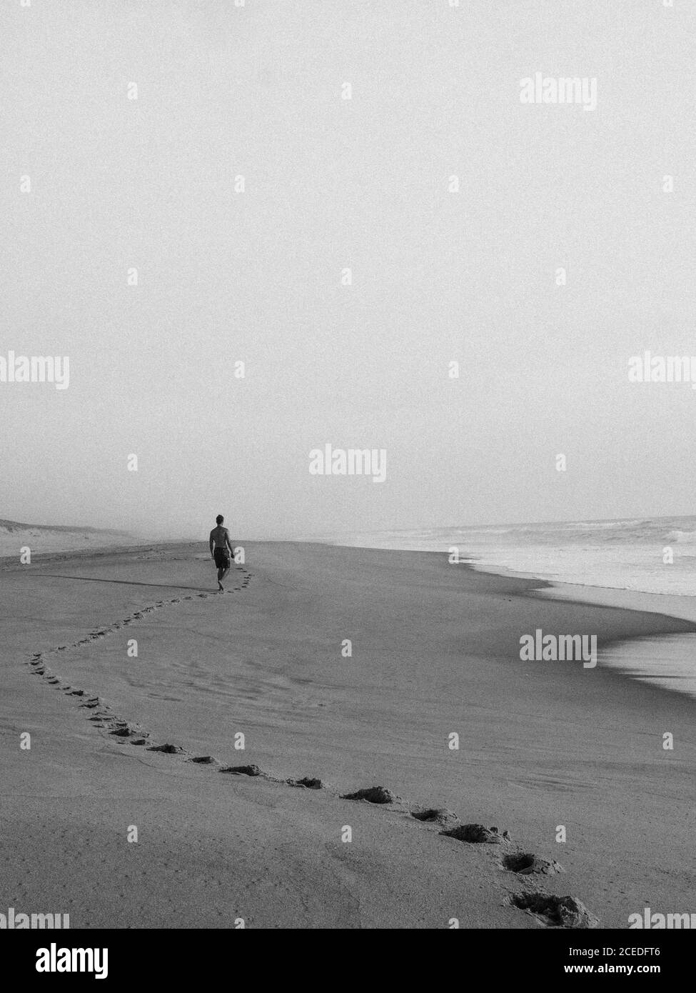Black and white shot of anonymous man walking on empty sandy coastline with huge ocean waves in haze Stock Photo