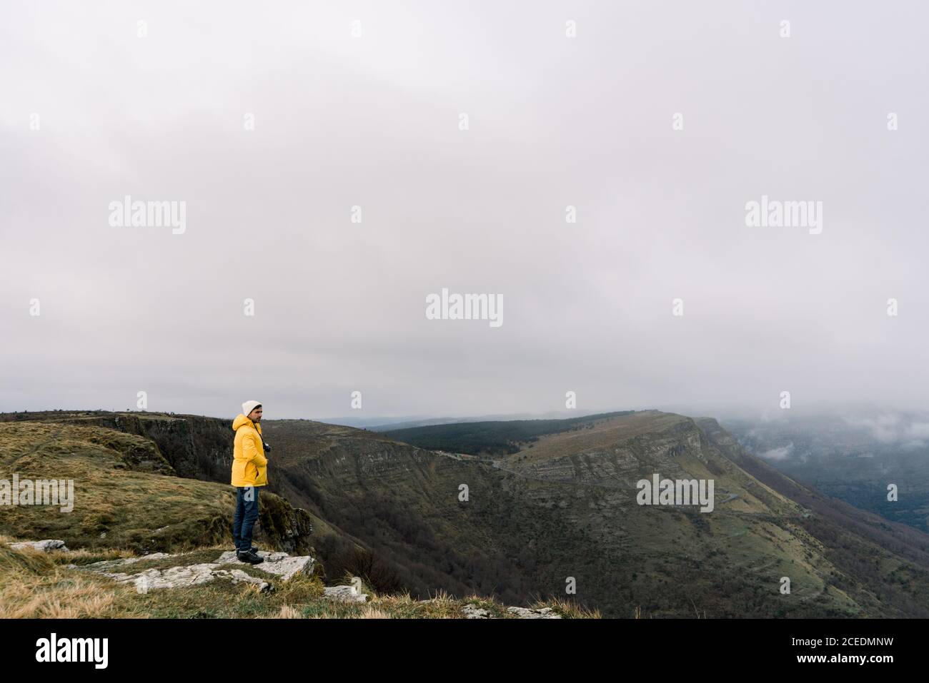 Side view of guy in yellow coat and hat standing on top of mountain and cloudy sky in Orduna, Spain Stock Photo