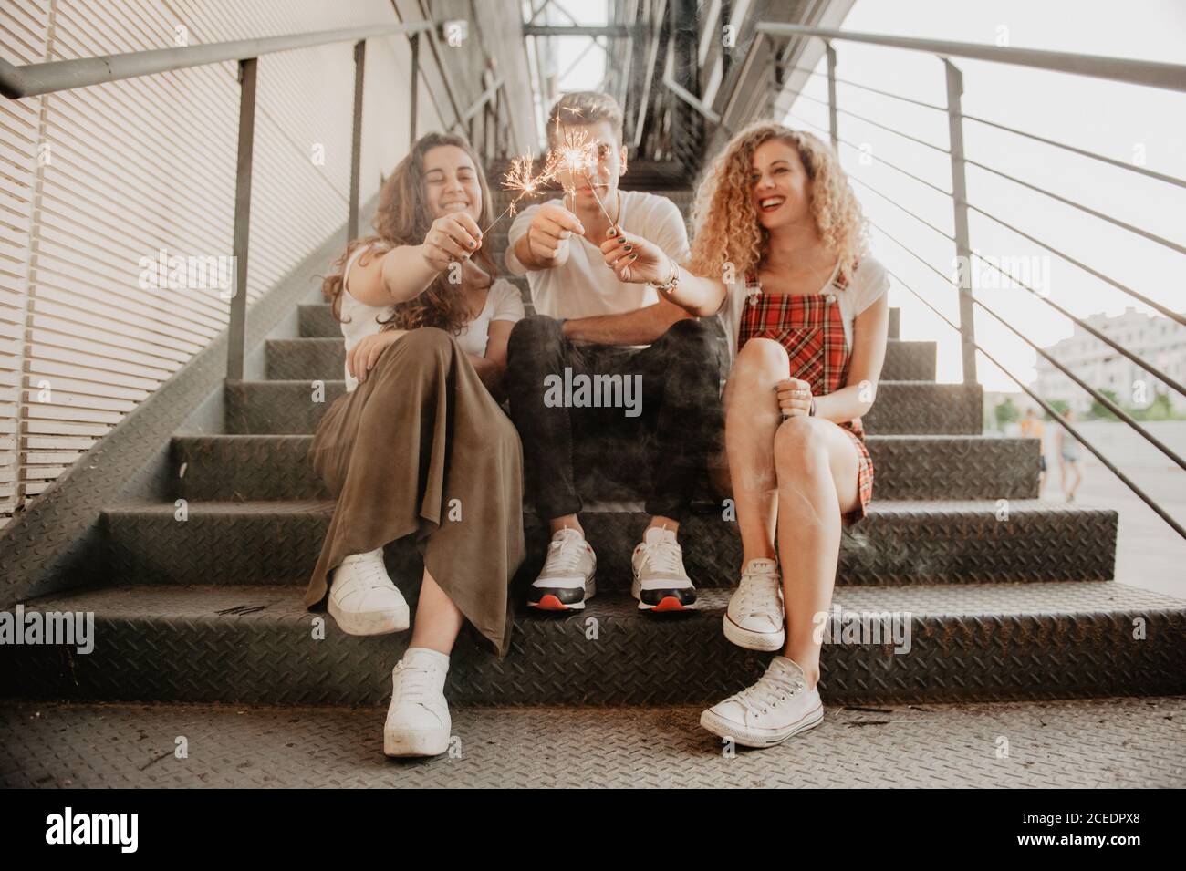 Three cheerful friends laughing and burning sparkles while sitting on metal stairs on city street Stock Photo
