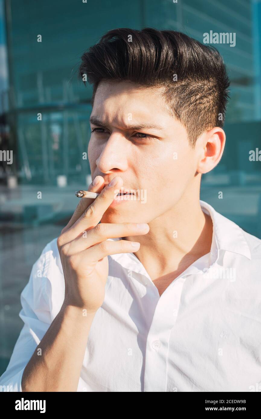 Young handsome with modern hairstyle wearing white shirt and smoking cigarette on street in sunlight Stock Photo