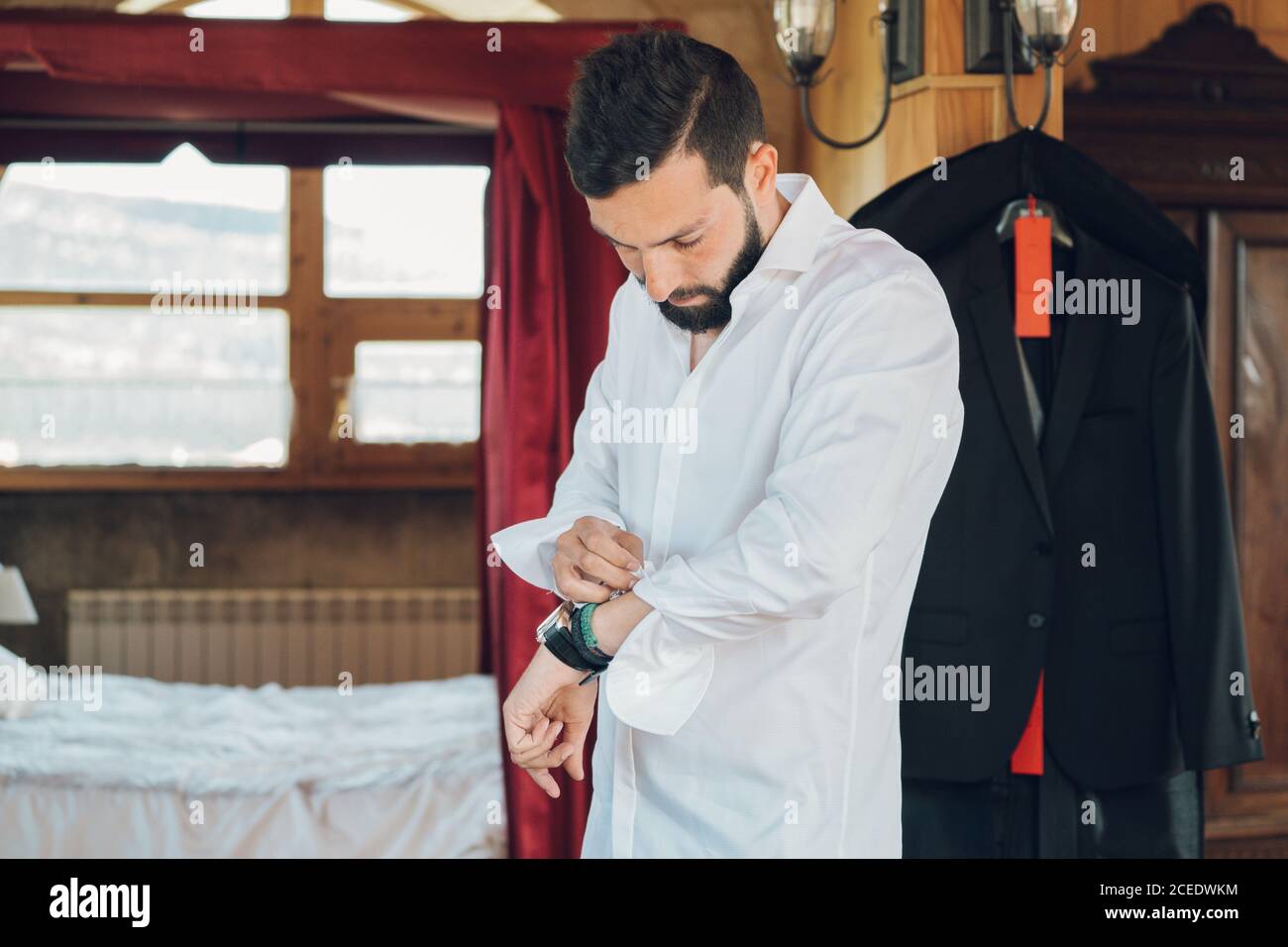 Young handsome man with dark hair and beard putting on white shirt in bedroom with new black costume hanging on clothes hanger Stock Photo